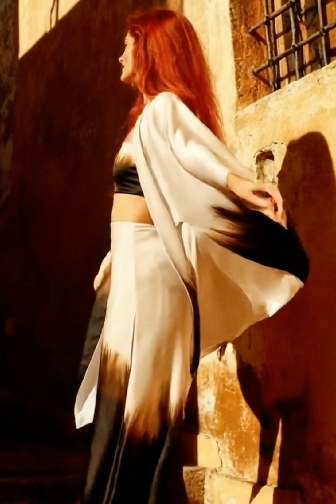 Woman with red hair draped in a white cloth against a textured wall.