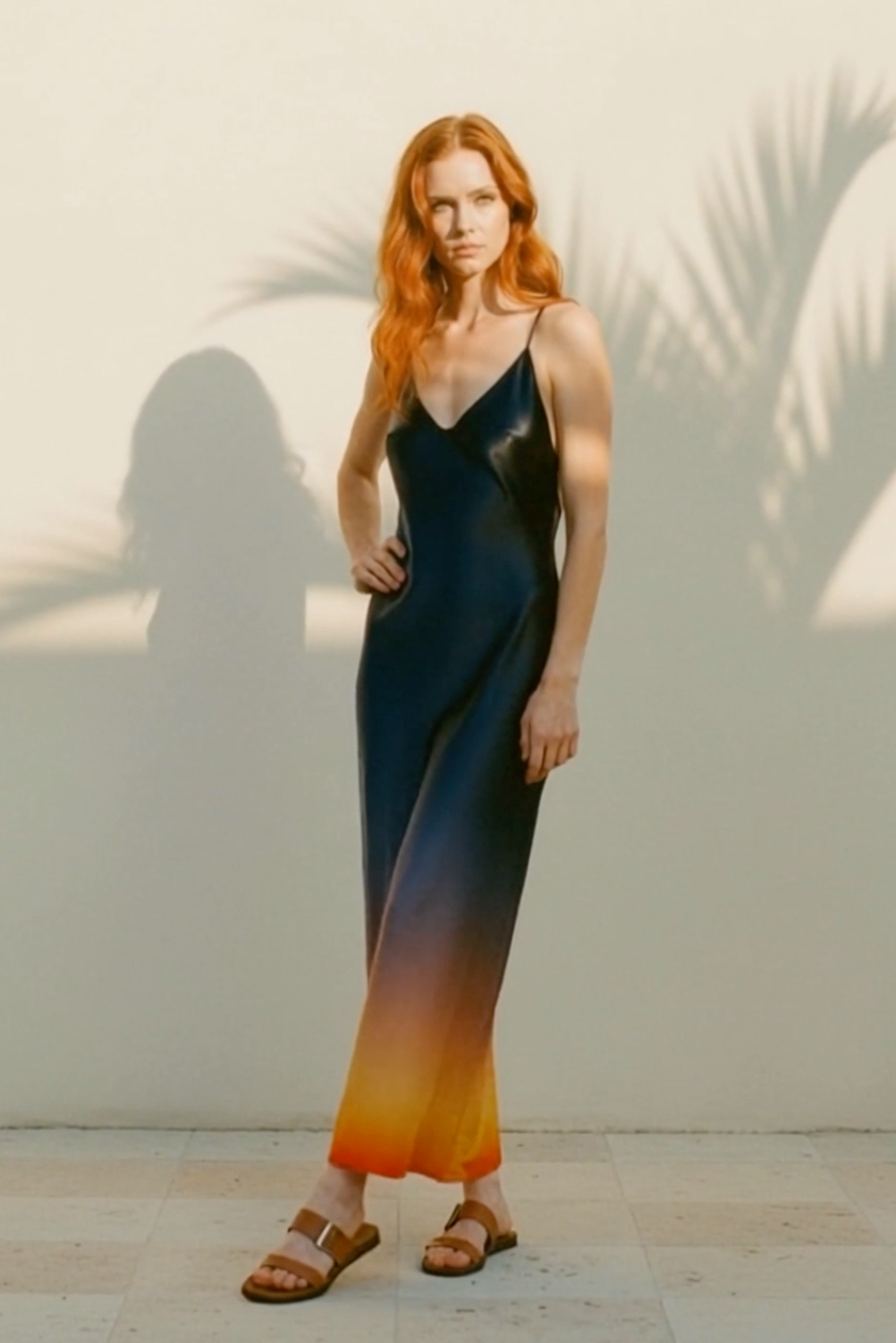 Woman in a gradient dress standing against a light wall with palm leaf shadows.