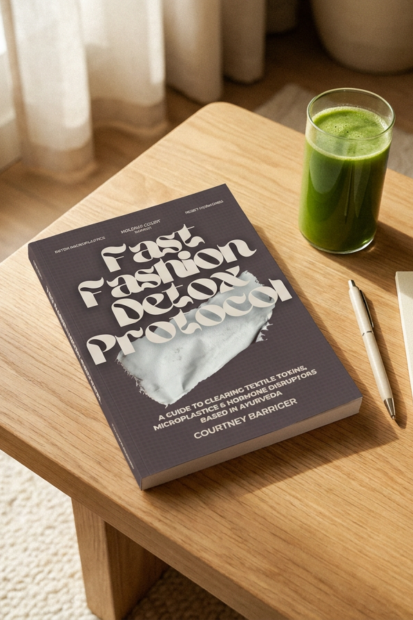 A book titled 'Fast Fashion Detox Protocol' by Courtney Bauermann lies on a light wood table next to a glass of green juice, a pen, and a small notebook in a bright, sunlit room.