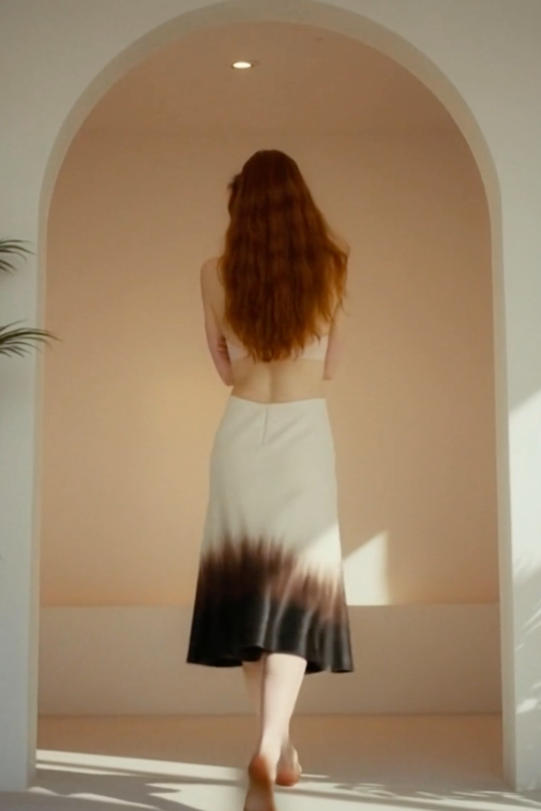 Woman in a gradient dress standing in a sunlit room with an archway.
