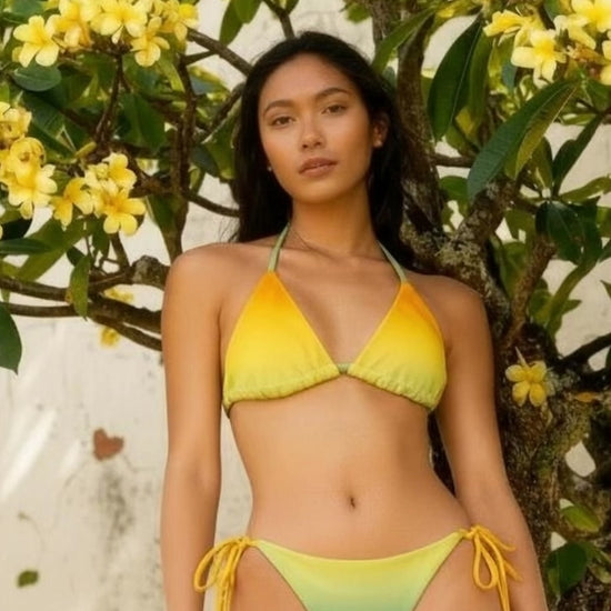 Vega String Bikini in Verde Horizons. Model wearing a yellow-to-green ombré string bikini under plumeria flowers, sustainable recycled swimwear, Holding Court Horizons collection.